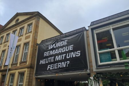 An der Fassade der Stadtbibliothek Osnabrück hängt ein schwarzes Banner mit weißer Aufschrift. Der Text fragt: Würde Remarque heute mit uns feiern?