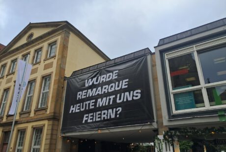 An der Fassade der Stadtbibliothek Osnabrück hängt ein schwarzes Banner mit weißer Aufschrift. Der Text fragt: Würde Remarque heute mit uns feiern?