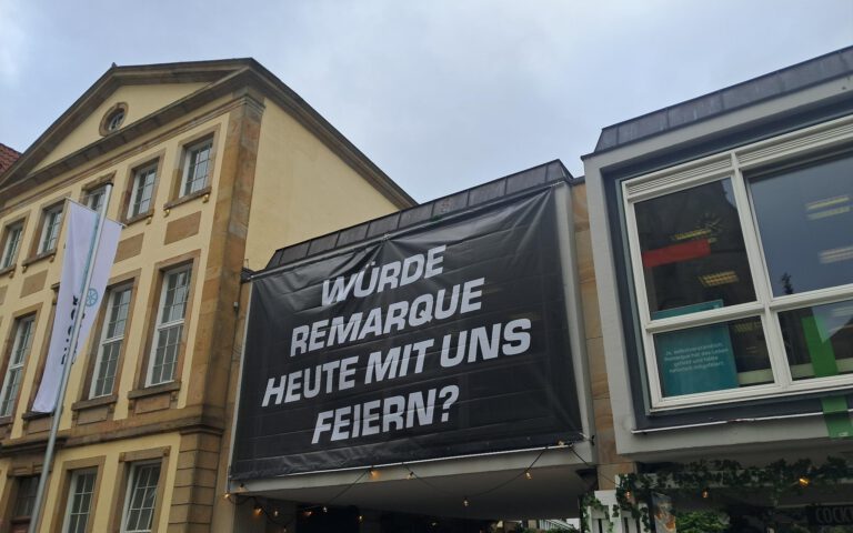 An der Fassade der Stadtbibliothek Osnabrück hängt ein schwarzes Banner mit weißer Aufschrift. Der Text fragt: Würde Remarque heute mit uns feiern?