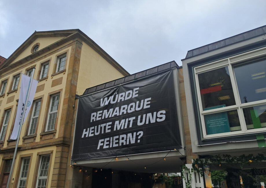 An der Fassade der Stadtbibliothek Osnabrück hängt ein schwarzes Banner mit weißer Aufschrift. Der Text fragt: Würde Remarque heute mit uns feiern?